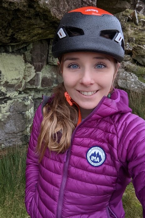 Photograph of Katie, this is a selfie, Katie is a white woman in her early thirties with bright blue eyes and naturally brown hair. In the photo she is wearing a purple thin down jacket with a badge that says IMA. She is also wearing a black and orange helmet.