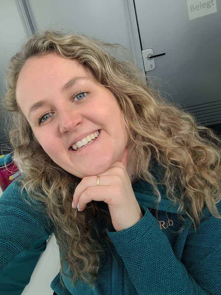 The photo is a selfie of Emily, she is a white woman in her later twenties, she has blonde-ish brown, curly hair. In the photo she is wearing a teal green jumper.